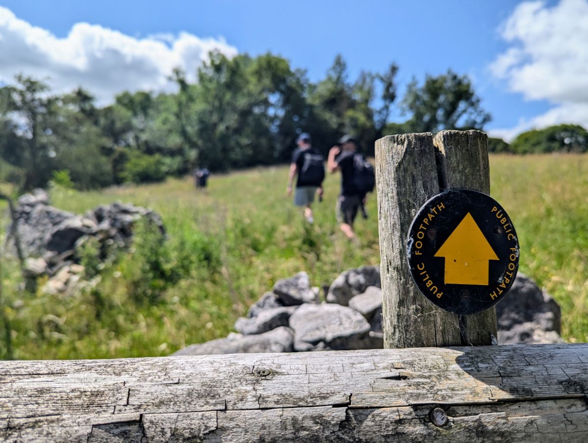 A UK footpath sign on a fencepost with a few walkers in the background.