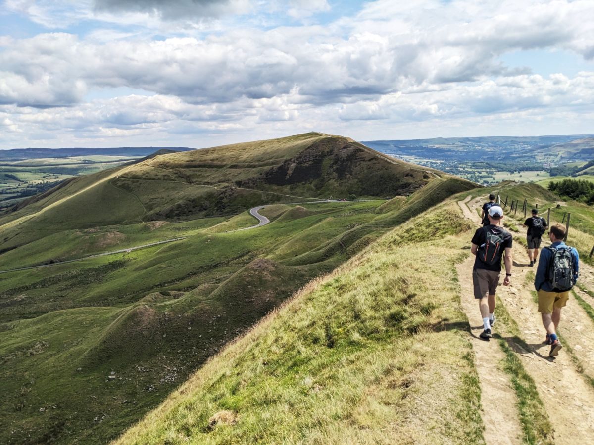 A few men walking along hills on a ridge under bright and cloud filled skies.